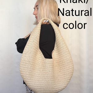 May include: A large, crescent-shaped hobo bag made of woven straw in a light khaki or natural color. The bag features a single integrated shoulder strap and a textured, braided surface. The text Khaki/Natural color is visible.