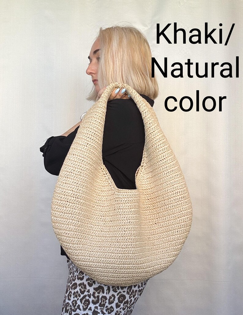May include: A large, crescent-shaped hobo bag made of woven straw in a light khaki or natural color. The bag features a single integrated shoulder strap and a textured, braided surface. The text Khaki/Natural color is visible.