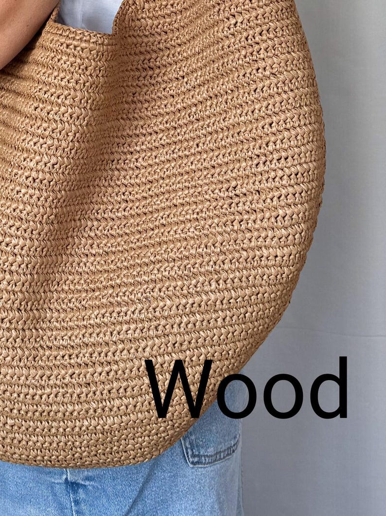 May include: A large, tan, woven straw tote bag with a textured, braided surface. The word Wood is printed in black, sans-serif capital letters in the lower right corner of the bag. The bag is positioned against a plain, light-colored background.