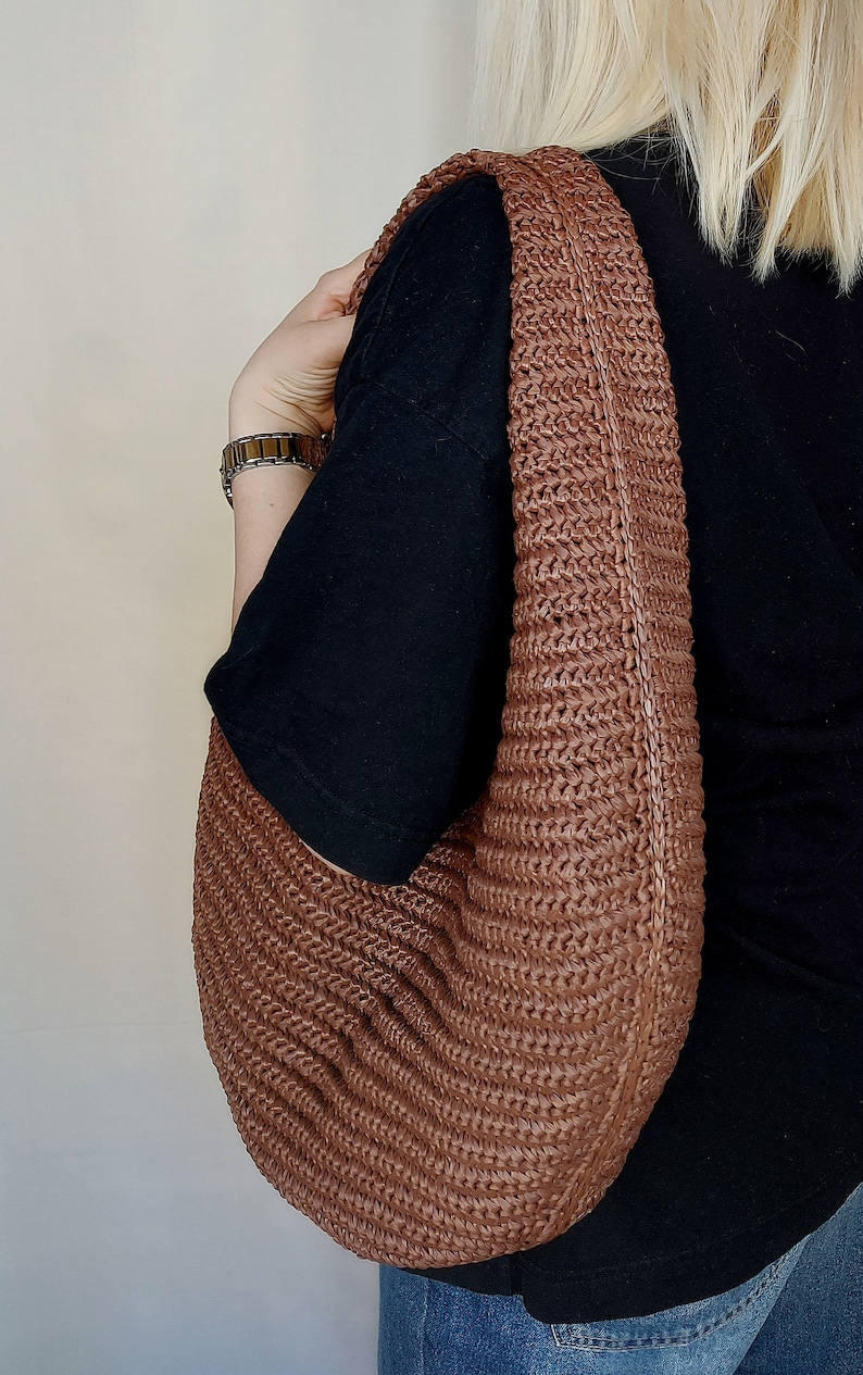 May include: A brown hobo-style shoulder bag crafted from a woven, textured material. The bag features a wide, integrated shoulder strap and a curved, slouchy silhouette with a consistent braided pattern throughout the entire exterior surface.