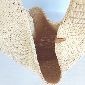 May include: A beige woven straw tote bag with an open top and a single integrated handle. The interior is visible, showing the textured weave of the material. A small tan leather tab with a metal rivet is attached to the upper edge of the bag.