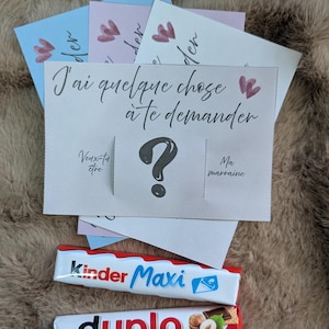 Puede incluir: Una pila de tarjetas rectangulares con fondos azul pastel, rosa y blanco, que presentan el texto en francés J'ai quelque chose à te demander, Veux-tu être y Ma marraine, con un signo de interrogación grande. Dos barritas de chocolate, Kinder Maxi y Duplo.