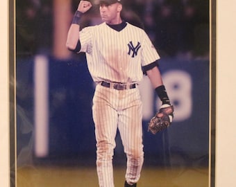 May include: A baseball player wearing a white and blue striped jersey with the New York Yankees logo, a black cap with the team's logo, and white pants with black stripes. The player is holding a baseball glove and raising his fist in the air.