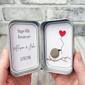 May include: A silver metal tin containing two white cards. The left card reads Happy 40th Anniversary, Alison & John, 12/05/1986. The right card features a small grey pebble bird perched on a brown branch, holding a red glittery heart balloon.