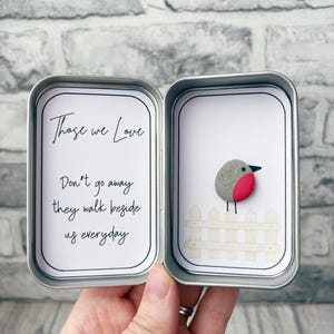May include: A silver metal tin containing two white cards. The left card features the black text: Those we Love Don't go away they walk beside us everyday. The right card displays a pebble art robin with a red breast perched on a white fence.