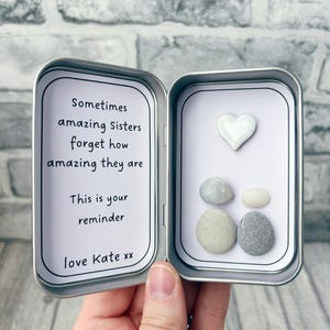 May include: A rectangular silver metal tin containing a white card with the printed text: Sometimes amazing Sisters forget how amazing they are. This is your reminder. love Kate xx. The right side features a white heart and four smooth pebbles.