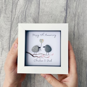 May include: A framed artwork with a white border. The artwork features two gray stone birds perched on a branch with pink flowers. A colorful flower is held by one bird. The text "Happy 15th Anniversary" and "Christina & Jack" are written on the artwork.