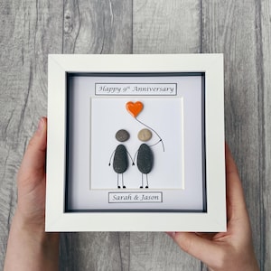 May include: A white framed picture with a white background. The picture shows two pebble people holding hands with a red heart balloon. The text "Happy 9th Anniversary" is at the top of the picture and "Sarah & Jason" is at the bottom.