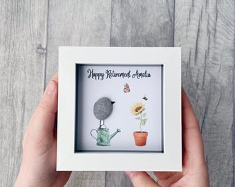 May include: A white framed picture with a gray stone bird perched on a green watering can. The picture also includes a sunflower with a butterfly flying above it. The text "Happy Retirement Amelia" is written on the picture.
