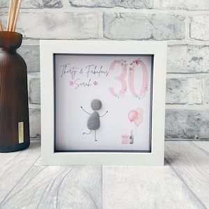 May include: A white framed picture with a pink background. The number 30 is written in white with pink flowers around it. The text "Thirty & Fabulous Sarah" is written in black. A grey pebble person is drawn with black marker, holding a balloon. There are two pink balloons and a gift box with a bottle of champagne.
