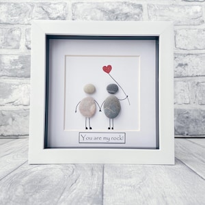 May include: A white framed picture with a hand-drawn image of two people made from stones holding a red heart balloon. The text "You are my rock!" is written below the image.