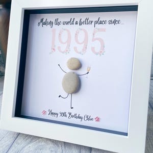May include: A white framed picture with a white background and black mat. The picture says "Making the world a better place since... 1995" with pink floral accents. A small pebble person is drawn with a glass of champagne. The bottom of the picture says "Happy 30th Birthday Chloe".