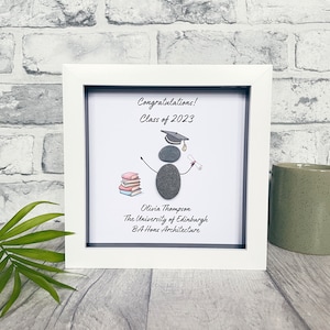 May include: A white framed picture with a graduation theme. The picture features a pebble person wearing a graduation cap and holding a diploma. The text reads "Congratulations! Class of 2023" and "Olivia Thompson, The University of Edinburgh, BA Hons Architecture".