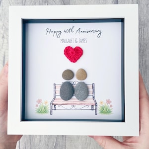 Peut inclure: Une œuvre d'art encadrée avec un cadre blanc. L'œuvre présente un cœur rouge, deux personnages en galets sur un banc et le texte "Happy 40th Anniversary" avec les noms "Margaret & James". Des fleurs décoratives sont de chaque côté du banc.