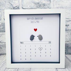 May include: A white framed wall art piece featuring a pebble art design of two birds and a red heart above a printed March 2026 calendar. The text reads Happy 40th Anniversary Julie & Mike, with a hand-drawn heart around the number 12.