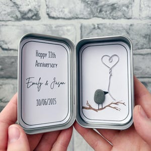 May include: A silver metal tin containing two white inserts. The left insert features the text Happy 11th Anniversary, Emily & Jason, and 10/06/2015. The right insert displays a small grey pebble bird on a painted branch holding a wire heart.