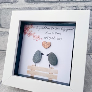 May include: White framed engagement art. The artwork shows two pebble birds on a wooden bench, a wooden heart with "Love", and the text "Congratulations On Your Engagement" with a date. The frame is square.