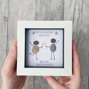 May include: A white framed pebble art piece. The artwork depicts two pebble figures toasting champagne glasses, with the text "HERE'S TO LOVE, LAUGHTER AND HAPPILY EVER AFTER." Below, it reads "ANNIE & JAMES ENGAGED 25TH OCTOBER 2025."