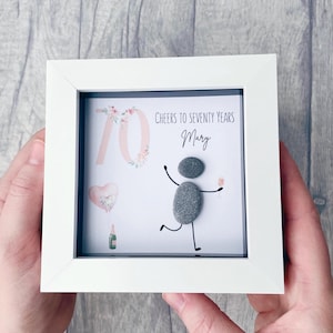 May include: A framed artwork with a white border. The artwork features a pebble art figure celebrating with a drink, a pink floral "70", a heart-shaped balloon, and the text "Cheers to Seventy Years Mary".
