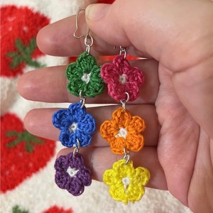 May include: Rainbow crochet flower earrings with silver hooks. Each earring features five crocheted flowers in different colors: green, pink, blue, purple, and yellow.