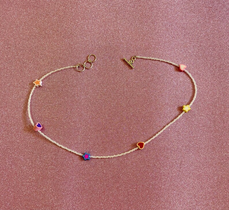 Op de afbeelding: Een witte kralenketting met kleurrijke hart- en sterbedels. De ketting heeft een zilveren sluiting en is perfect om elke outfit een vleugje speelsheid te geven.