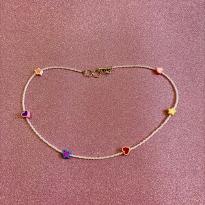 Op de afbeelding: Een witte kralenketting met kleurrijke hart- en sterbedels. De ketting heeft een gouden sluiting.