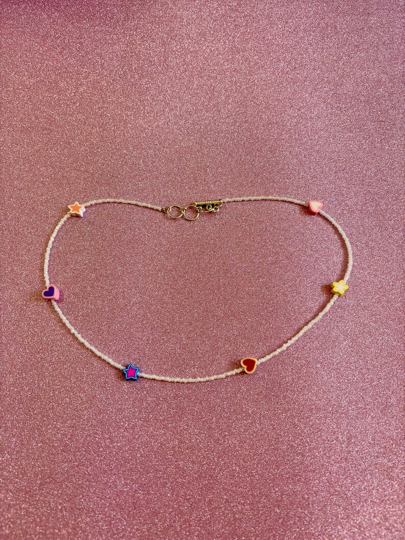 Op de afbeelding: Een witte kralenketting met kleurrijke hart- en sterbedels. De ketting heeft een gouden sluiting.