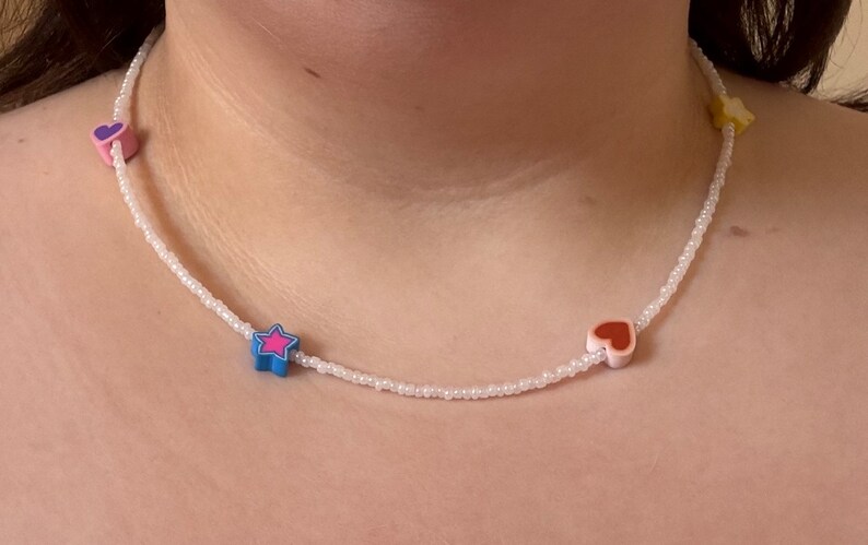 Op de afbeelding: Een witte kralenketting met kleurrijke hart- en sterbedels. De ketting is gemaakt van kleine witte kralen en heeft een roze hart, een rood hart, een blauwe ster met een roze centrum en een gele ster.