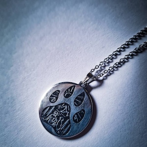 Puede incluir: Un collar de plata con un colgante redondo que presenta un diseño grabado detallado de una huella de pata con una escena de fogata en su interior.