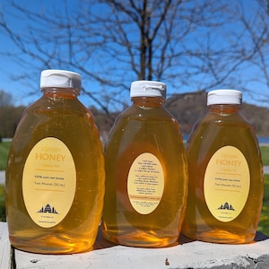 May include: Three clear plastic bottles of honey labeled "Upstate Honey" with a white label and a white cap. The bottles are sitting on a wooden surface. The label includes the text "100% pure raw honey" and "Two Pounds (32 oz.)".