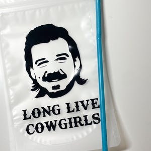 Op de afbeelding: Een doorzichtige plastic drinkzak met een zwarte grafische illustratie van een persoon met een snor en een matje, met de tekst LONG LIVE COWGIRLS eronder gedrukt. Een lang, dun, felblauw plastic rietje ligt naast de drinkzak.