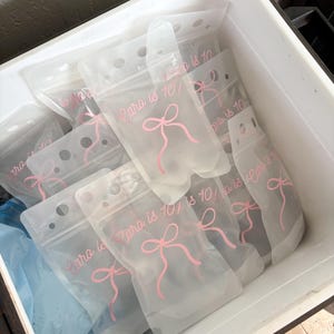 Op de afbeelding: Een verzameling doorschijnende, flexibele plastic drinkzakjes met een hersluitbare bovenkant. Elk zakje is voorzien van een roze gedrukte afbeelding van een strik en de tekst "Cara is 10!" in een cursief lettertype. De zakjes liggen in een witte bak.