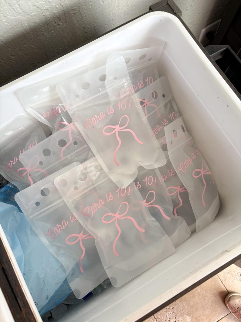 Op de afbeelding: Een verzameling doorschijnende, flexibele plastic drinkzakjes met een hersluitbare bovenkant. Elk zakje is voorzien van een roze gedrukte afbeelding van een strik en de tekst "Cara is 10!" in een cursief lettertype. De zakjes liggen in een witte bak.