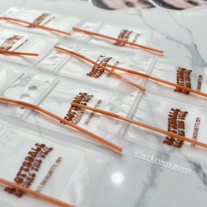 Op de afbeelding: Een verzameling doorzichtige plastic zakjes met oranje rietjes, elk bedrukt met de tekst Scottsdale Before The Veil en Suli's Bachelorette 2024, vergezeld door verschillende ronde papieren uitsparingen met een mannengezicht op stokjes.