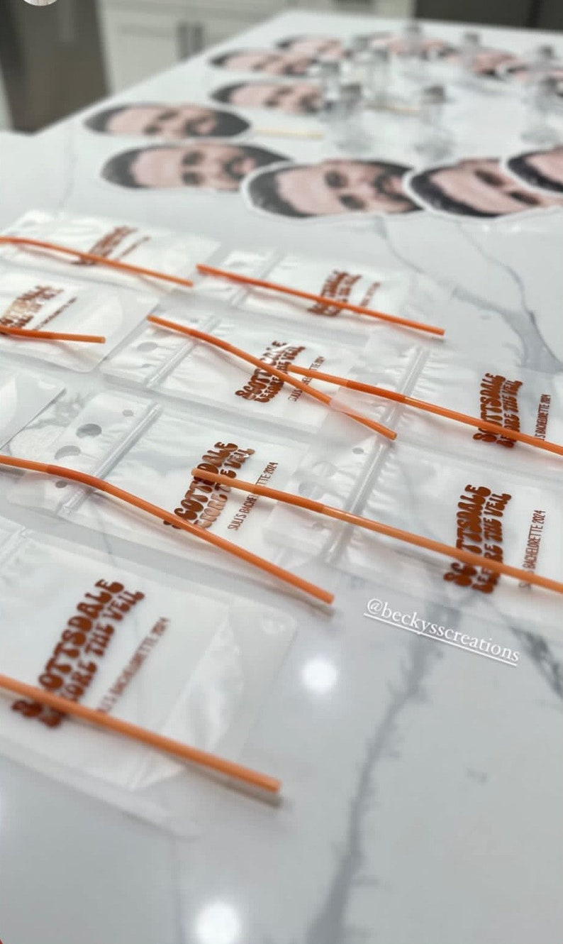 Op de afbeelding: Een verzameling doorzichtige plastic zakjes met oranje rietjes, elk bedrukt met de tekst Scottsdale Before The Veil en Suli's Bachelorette 2024, vergezeld door verschillende ronde papieren uitsparingen met een mannengezicht op stokjes.