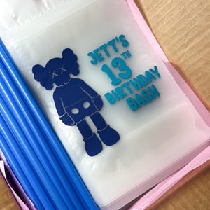 Op de afbeelding: Een stapel doorzichtige plastic drinkzakjes met blauwe rietjes. Elk zakje is voorzien van een donkerblauwe afbeelding van een stripfiguur met X-vormige ogen en de tekst JETT'S 13TH BIRTHDAY BASH in het blauw gedrukt.