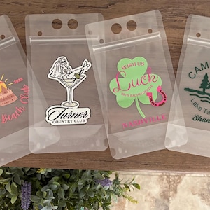 Op de afbeelding: Vier doorzichtige, flexibele plastic drinkzakjes met ritssluiting en handvatgaten. Elk zakje heeft een unieke afbeelding: een strandsc&egrave;ne met 'Josie's Beach Club', een persoon in een martiniglas met 'Turner Country Club', een klavertje met 'Wish Us Luck' en een bossc&egrave;ne met 'Camp Bach Lake Tahoe, CA'.