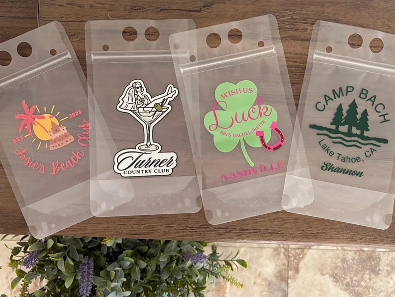 Op de afbeelding: Vier doorzichtige, flexibele plastic drinkzakjes met ritssluiting en handvatgaten. Elk zakje heeft een unieke afbeelding: een strandsc&egrave;ne met 'Josie's Beach Club', een persoon in een martiniglas met 'Turner Country Club', een klavertje met 'Wish Us Luck' en een bossc&egrave;ne met 'Camp Bach Lake Tahoe, CA'.