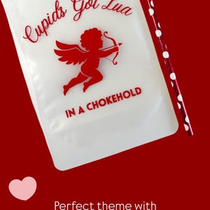 Op de afbeelding: Een doorschijnend drinkzakje met een rode afbeelding van een cherubijn met pijl en boog. De tekst luidt "Cupid's Got Lua" in cursief en "IN A CHOKEHOLD" in blokletters. Een rood-wit gestippeld rietje ligt naast het drinkzakje.