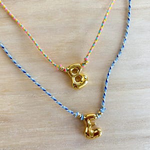 May include: Two gold-colored initial necklaces. One necklace has a gold letter "B" pendant, and the other has a gold letter "E" pendant. The necklaces have colorful braided cords in blue, pink, yellow, and gold.