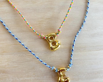Peut inclure: Deux colliers avec des initiales dorées. L'un a un pendentif en forme de lettre "B" dorée, et l'autre a un pendentif en forme de lettre "E" dorée. Les colliers ont des cordons tressés colorés en bleu, rose, jaune et or.