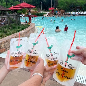 Op de afbeelding: Vier doorzichtige plastic drinkzakjes gevuld met een amberkleurige vloeistof, elk met een rood rietje en een groene afbeelding van een cocktailglas met muizenoortjes. De tekst "We've Got Ears Say Cheers" is in het zwart op elk zakje gedrukt.