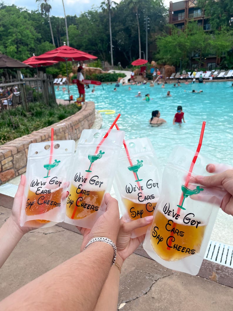 Op de afbeelding: Vier doorzichtige plastic drinkzakjes gevuld met een amberkleurige vloeistof, elk met een rood rietje en een groene afbeelding van een cocktailglas met muizenoortjes. De tekst "We've Got Ears Say Cheers" is in het zwart op elk zakje gedrukt.