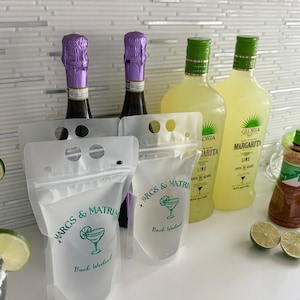 Op de afbeelding: Twee doorschijnende plastic drankzakjes met een ritssluiting en handvatgaten, bedrukt met groene tekst MARGS & MATRIMONY en Bach Weekend naast een margaritaglas-icoon, geplaatst voor twee wijnflessen en twee margaritaflessen.