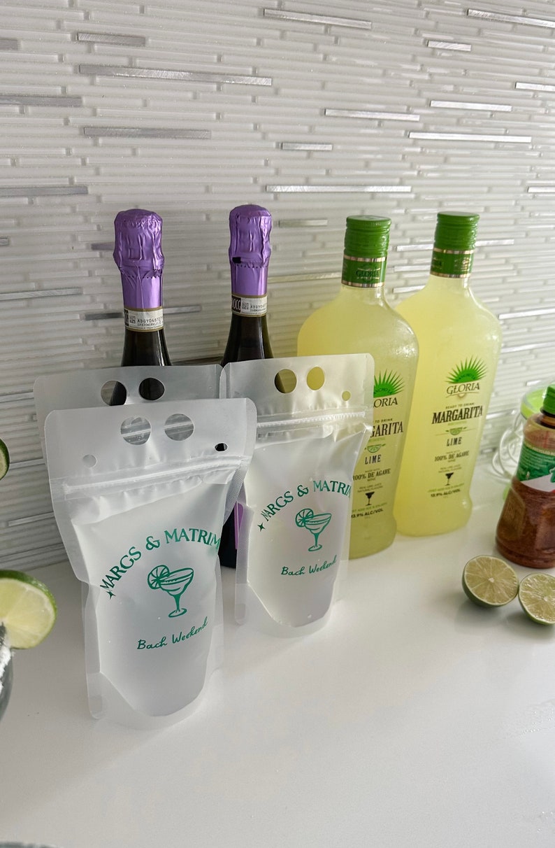 Op de afbeelding: Twee doorschijnende plastic drankzakjes met een ritssluiting en handvatgaten, bedrukt met groene tekst MARGS & MATRIMONY en Bach Weekend naast een margaritaglas-icoon, geplaatst voor twee wijnflessen en twee margaritaflessen.