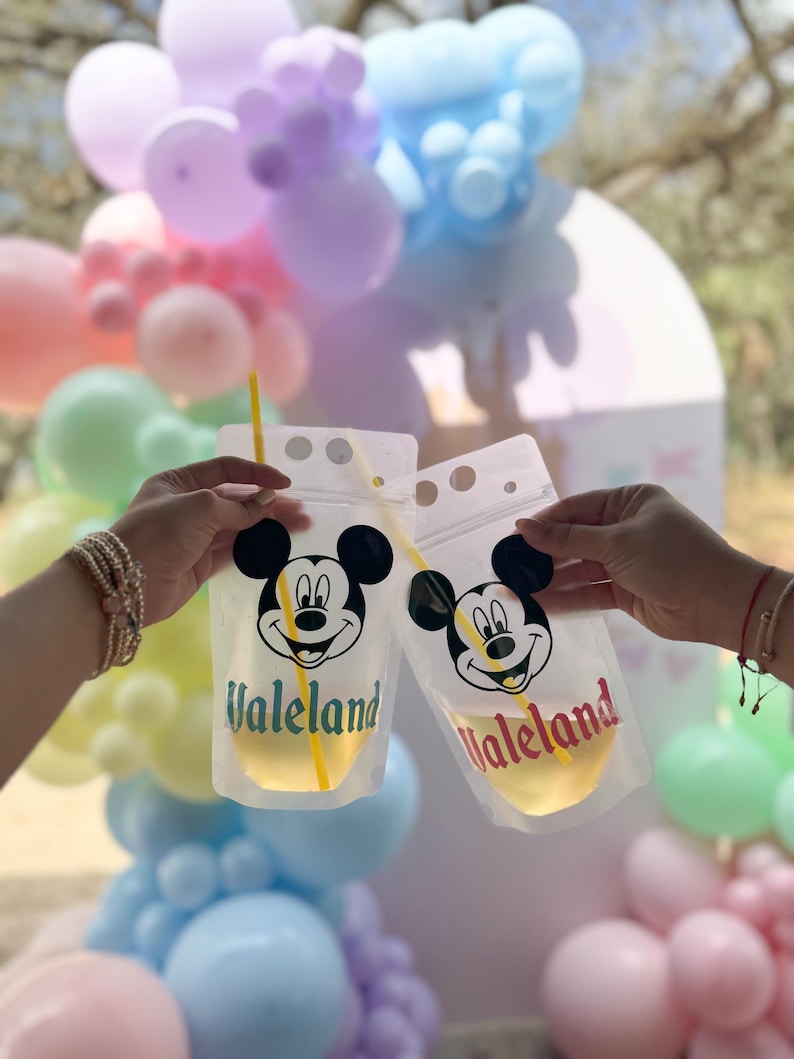 Op de afbeelding: Twee doorzichtige plastic drinkzakjes met een zwart-witte Mickey Mouse-afbeelding en de tekst Valeland in respectievelijk groen en rood. Elk zakje bevat een geel rietje en een lichtgekleurde vloeistof, gehouden voor een achtergrond van pastelballonnen.