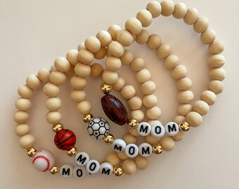 May include: Three wooden bracelets with gold beads and charms. The charms include a baseball, basketball, soccer ball, and a football. The bracelets also have the word "MOM" spelled out on them.