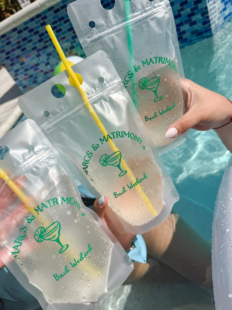 Op de afbeelding: Drie doorzichtige, flexibele plastic drinkzakjes met gele rietjes, elk bedrukt met een groen margaritaglas-icoon en de tekst "MARGS & MATRIMONY" boven "Bach Weekend". De zakjes zijn gevuld met een lichtgekleurde vloeistof en vertonen condens.