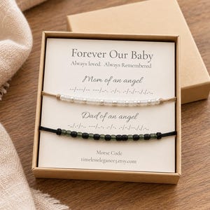May include: Two Morse code bracelets in a gift box. One bracelet has white beads on a tan cord labeled Mom of an angel. The other has dark green beads on a black cord labeled Dad of an angel. The card reads Forever Our Baby, Always loved. Always Remembered.