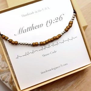 May include: A beaded necklace with brown and silver beads, presented on a card inside a box. The card displays the text "Matthew 19:26" and Morse code. The words "Handmade in the U.S.A." are also visible.